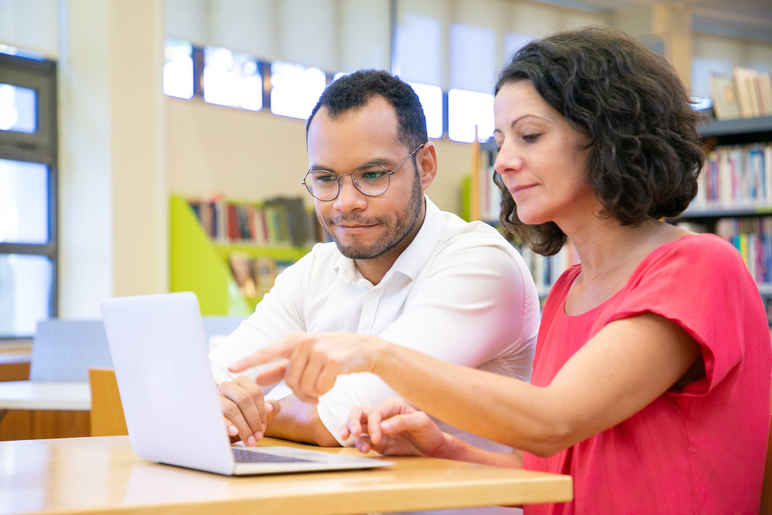 Positive adult students doing academic research in library. Man and woman in casual sitting at desk, using laptop, pointing at display, watching content. College concept